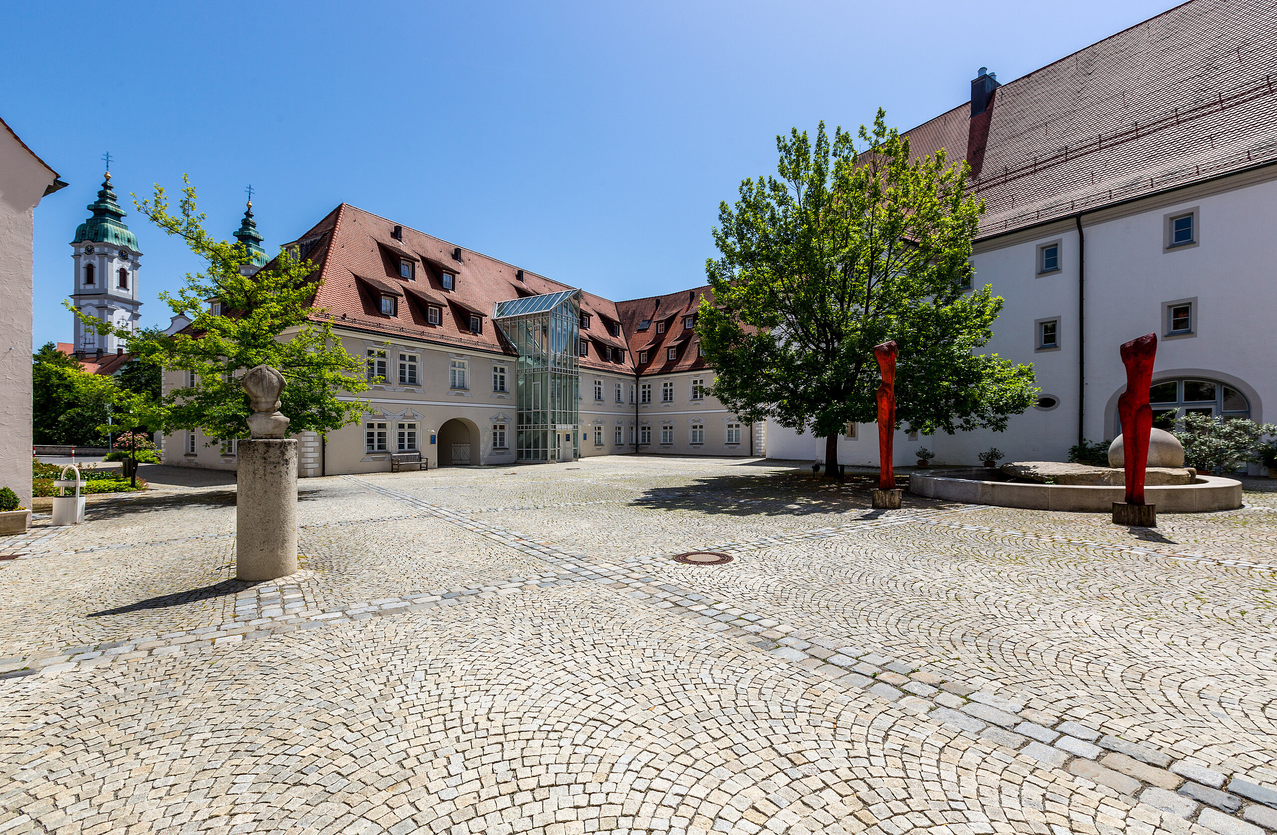 Gepflasterter Innenhof der Klinik im Hofgarten in Bad Waldsee, umgeben von historischen Gebäuden mit roten Dächern, Bäumen und blauem Himmel im Hintergrund