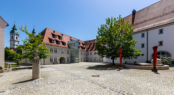 Gepflasterter Innenhof der Klinik im Hofgarten in Bad Waldsee, umgeben von historischen Gebäuden mit roten Dächern, Bäumen und blauem Himmel im Hintergrund