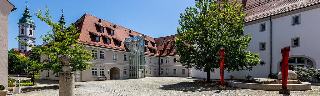 Gepflasterter Innenhof der Klinik im Hofgarten in Bad Waldsee, umgeben von historischen Gebäuden mit roten Dächern, Bäumen und blauem Himmel im Hintergrund