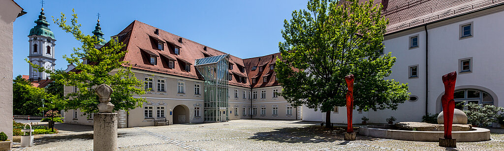 Gepflasterter Innenhof der Klinik im Hofgarten in Bad Waldsee, umgeben von historischen Gebäuden mit roten Dächern, Bäumen und blauem Himmel im Hintergrund